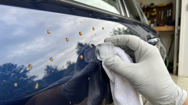 Main gantée frottant de la résine de carrosserie sur une voiture bleue avec un glaçon et un chiffon blanc.