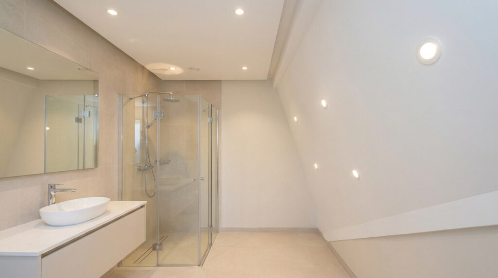 Salle de bain lumineuse avec spots LED encastrés au plafond et dans le mur incliné au-dessus de la douche.