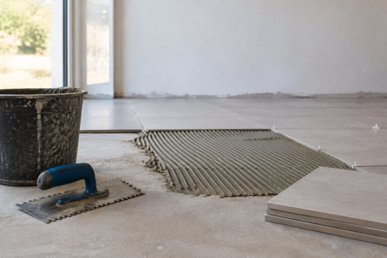 Un chantier de carrelage avec une taloche, un seau, de la colle peignée et des carreaux gris sur un sol en béton.