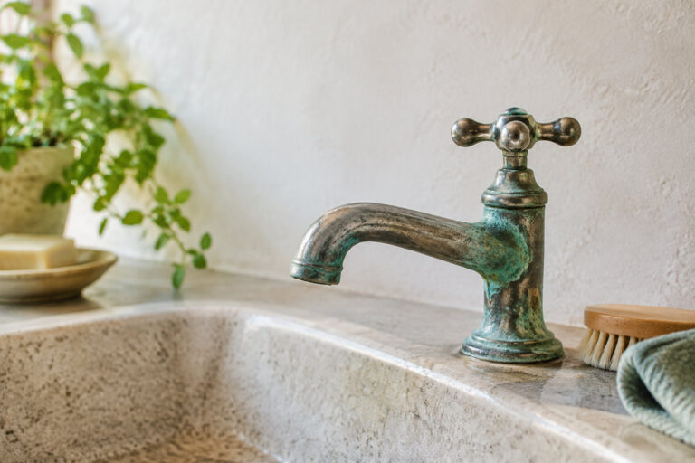 Un robinet vintage en métal avec des traces de vert-de-gris posé sur un évier en pierre dans une salle de bain lumineuse.