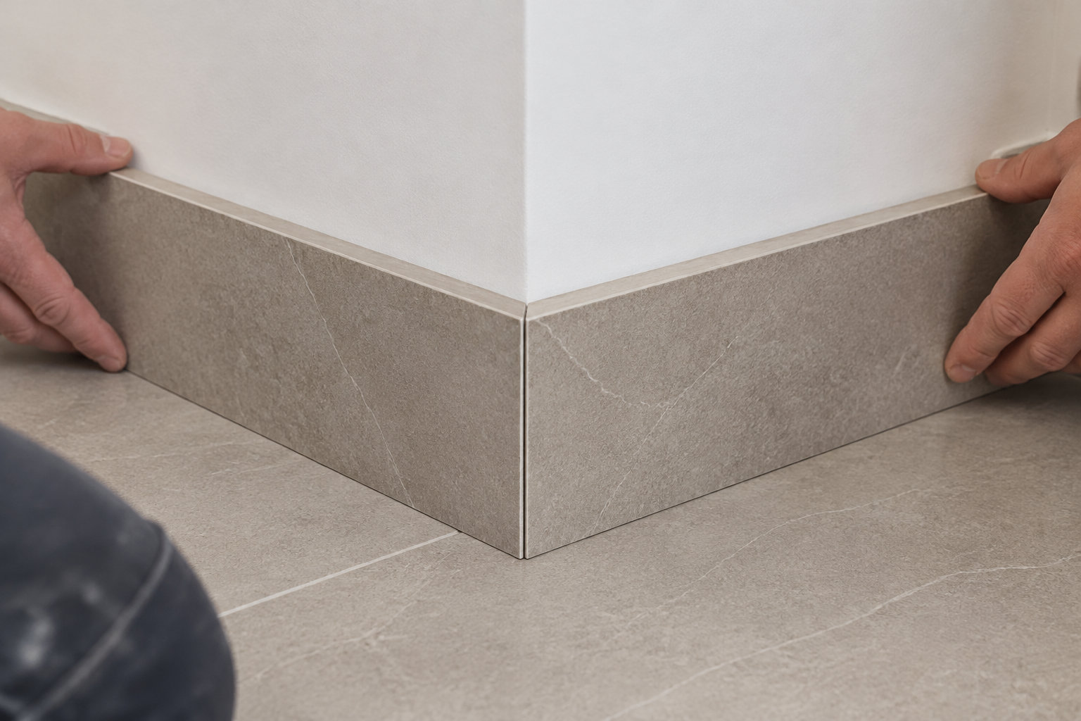 Deux mains ajustent des plinthes en carrelage gris au pied d’un mur blanc, sur un sol carrelé.